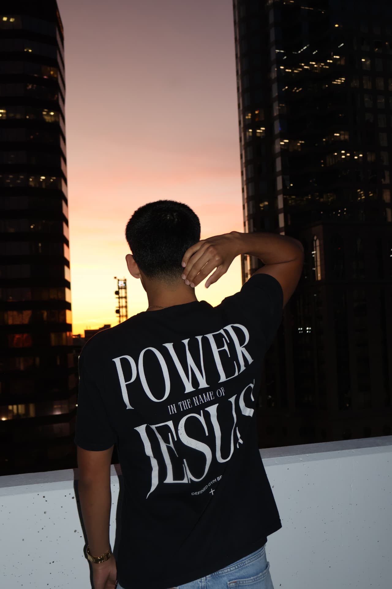 Model wearing the Power In The Name Of Jesus tee against a city skyline at dusk