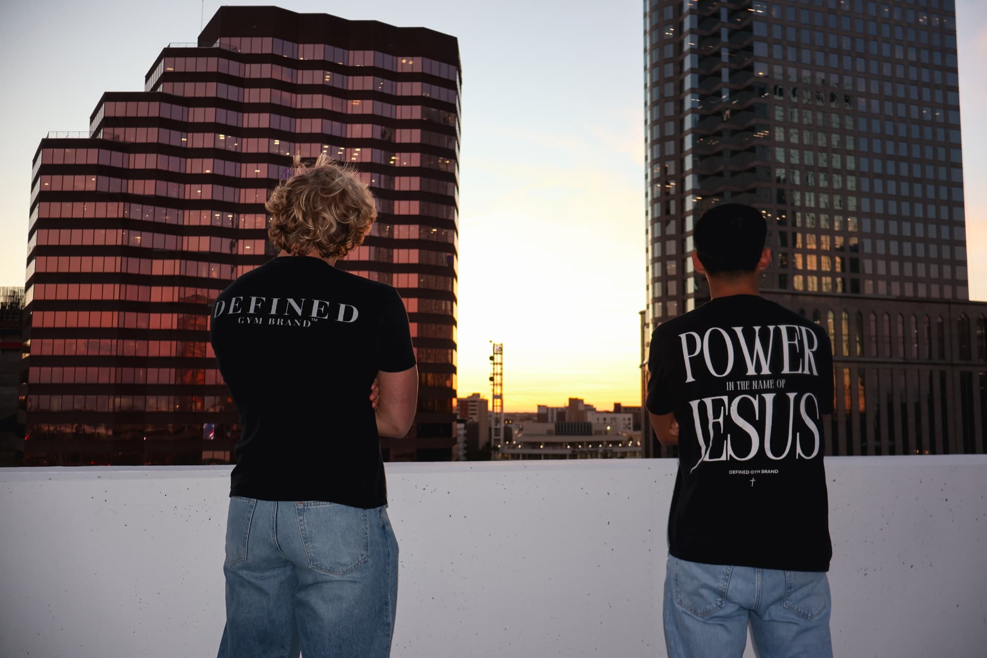 Two models on a rooftop at sunset wearing Defined Gym Brand and Power In The Name Of Jesus tees